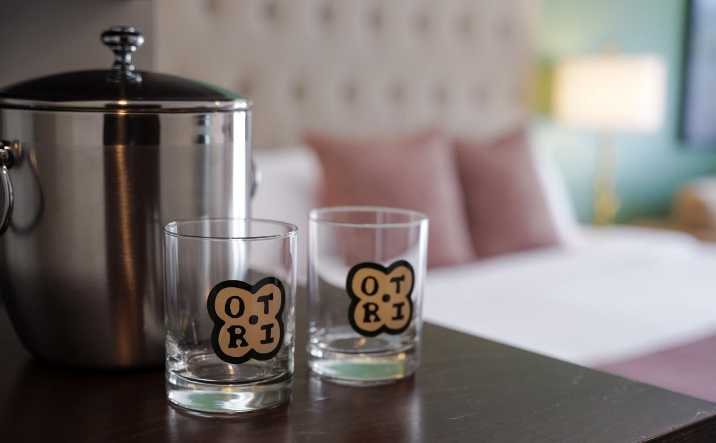 Two branded glasses and an ice bucket on a table against a blurred hotel room backdrop.