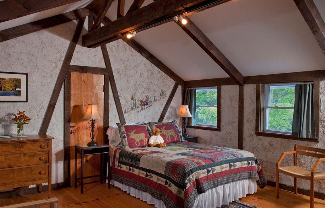 Cozy attic bedroom with wooden beams, quilted bed, and two windows.