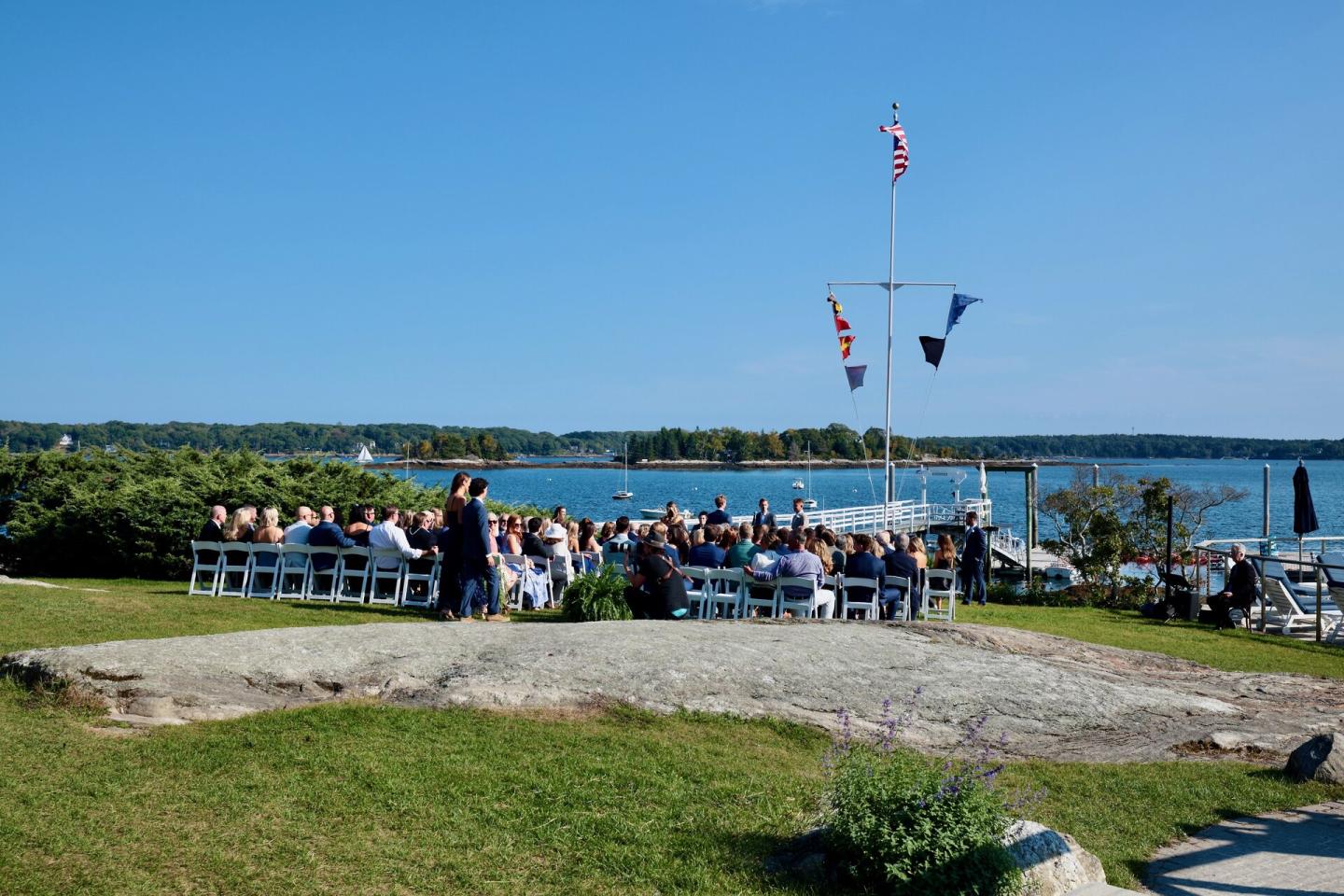 Outdoor wedding by the sea with guests seated on white chairs under a clear blue sky.OUtdoor Cer