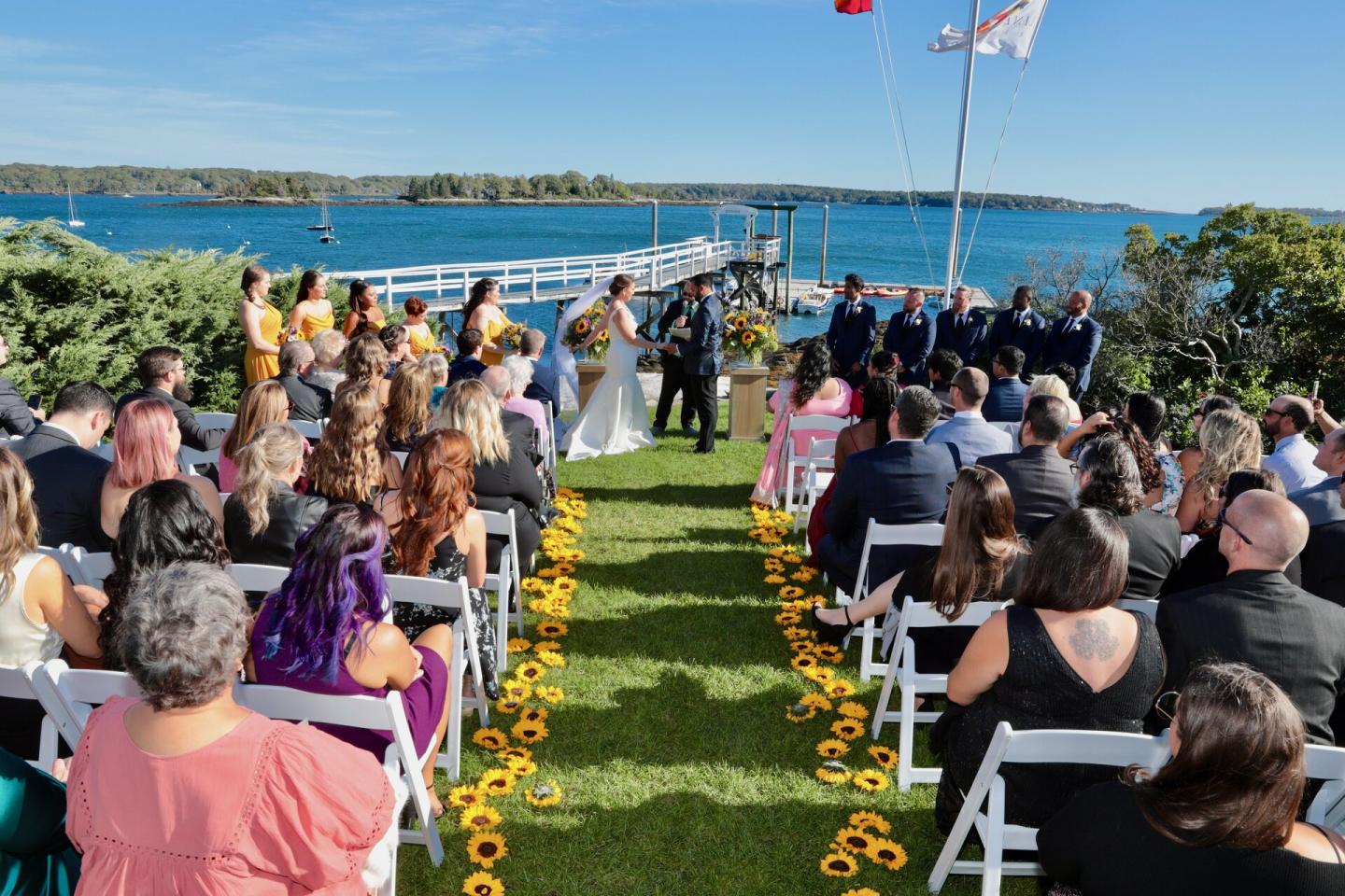 Outdoor wedding by a lake, guests seated, couple at the altar under a sunny sky.