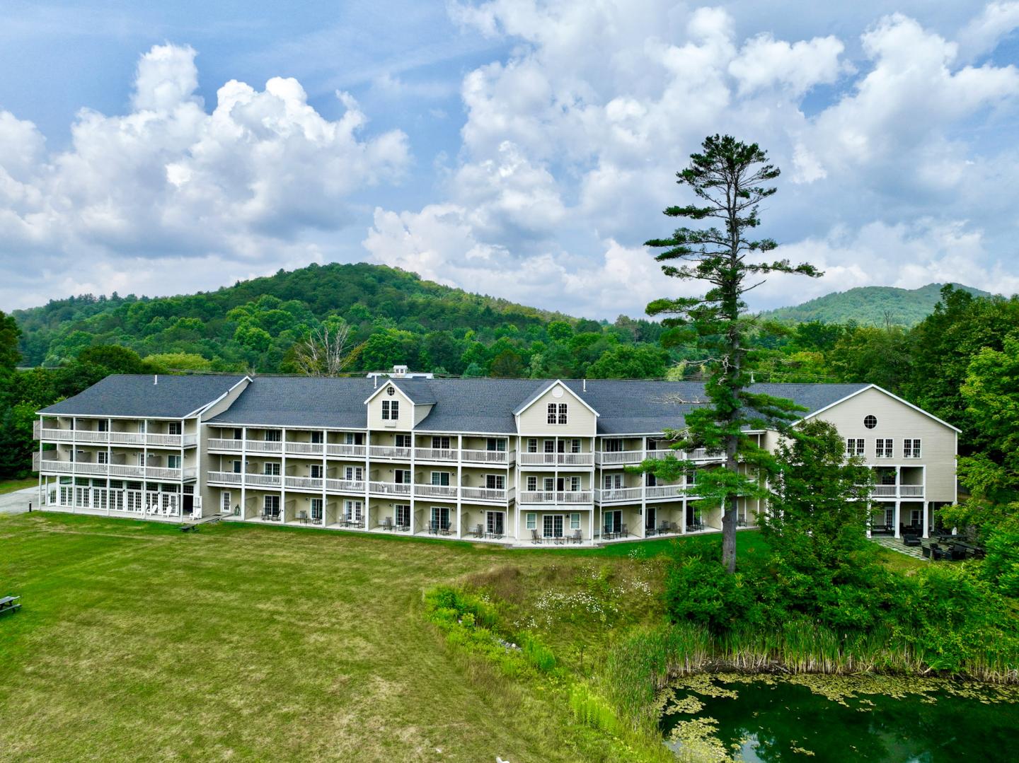 Large white building with balconies, set against a green hill and cloudy blue sky.