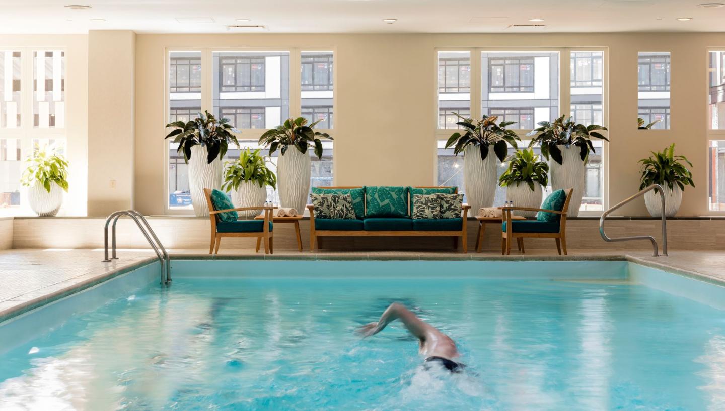 Indoor pool with swimmer, potted plants, and seating area. Bright windows background.