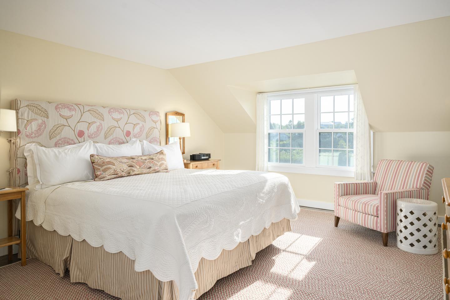 Cozy bedroom with a double bed, floral armchair, and soft natural light from a window.