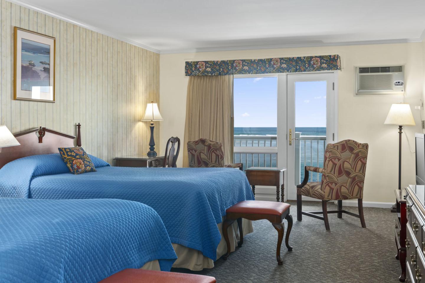 Hotel room with two blue-covered beds, lamps, and ocean view.