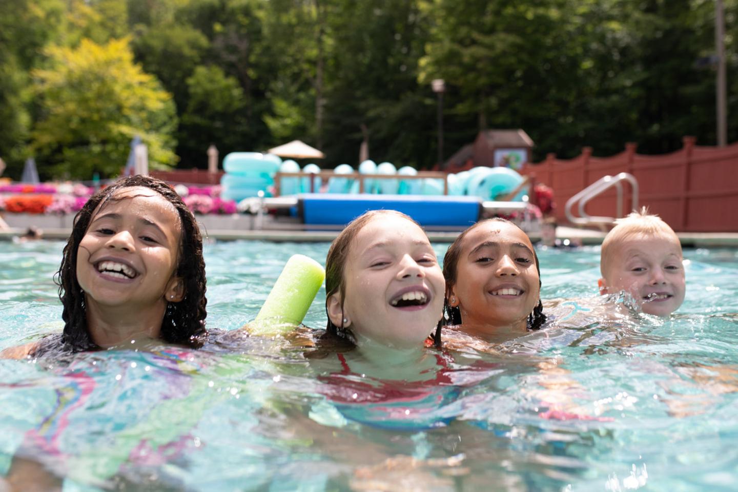 Four kids smiling while swimming in a pool on a sunny day.