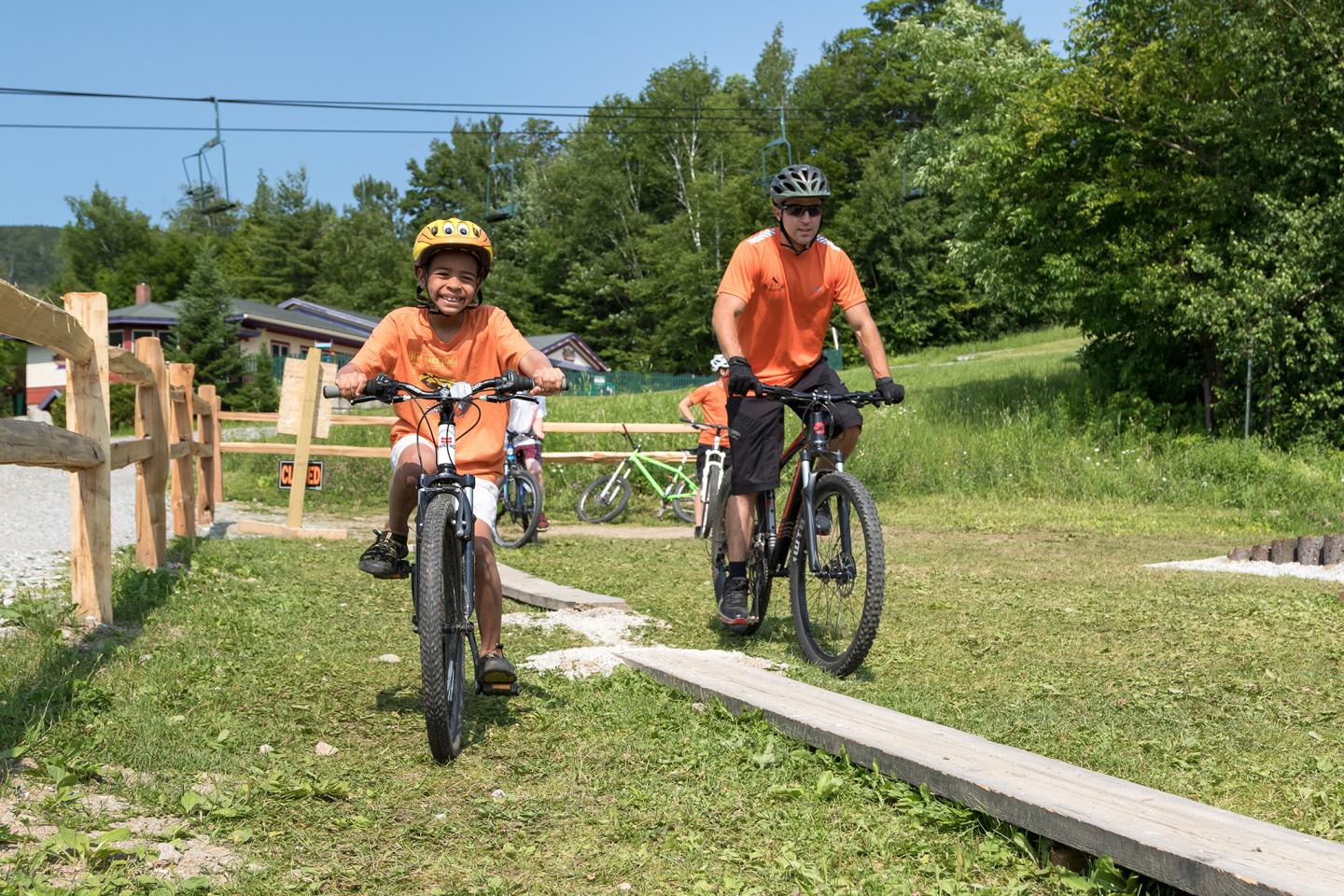 Children biking on grass trail, wearing helmets, under a sunny sky.