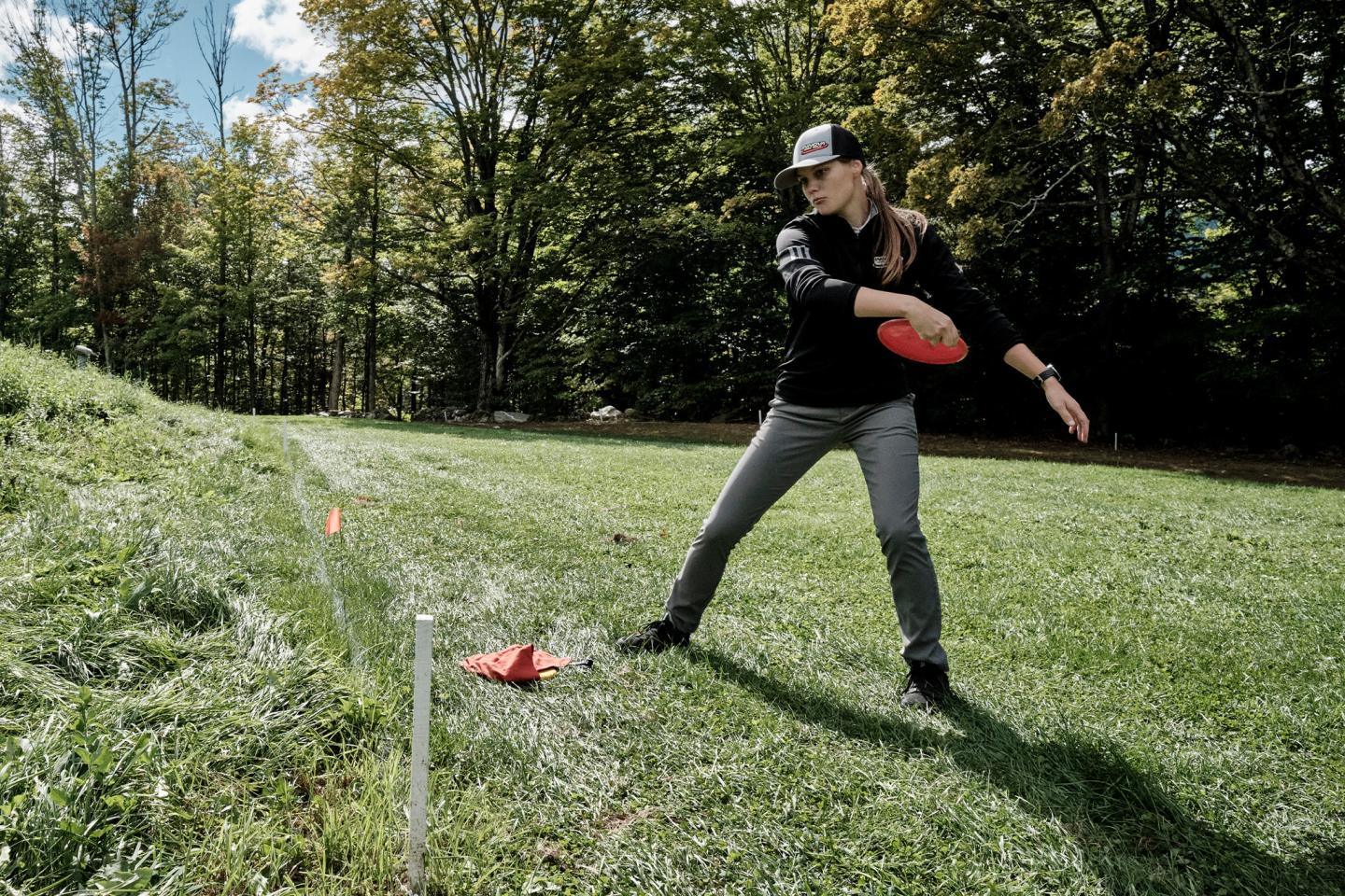 Woman throwing a frisbee in a grassy field, surrounded by trees.