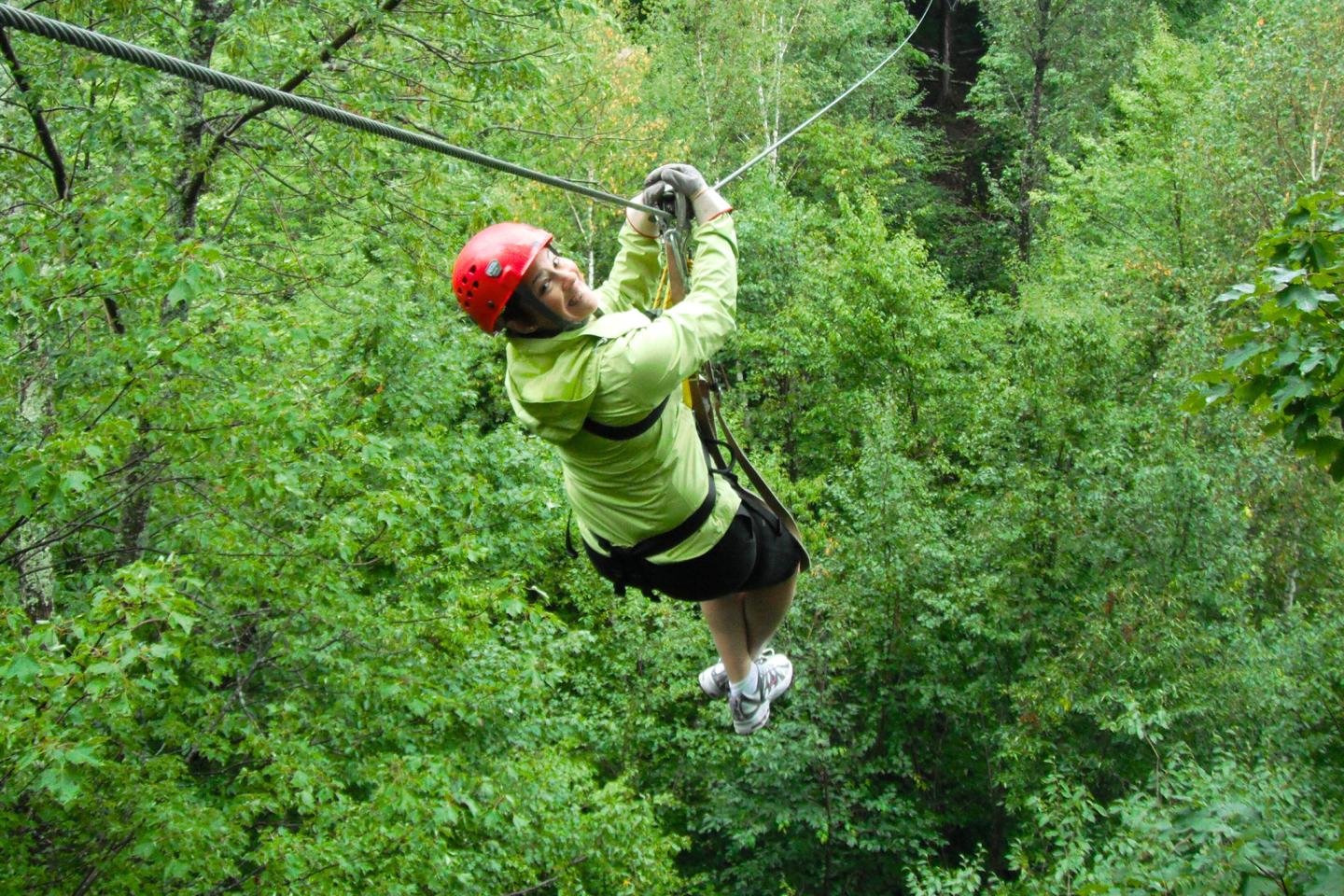 Person ziplining through lush green forest, wearing red helmet and green jacket.