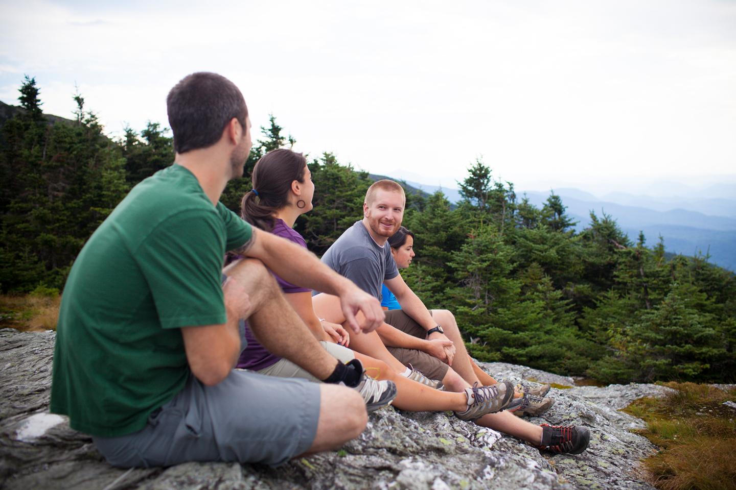 Three people sitting on a rocky hill, overlooking trees and distant mountains.