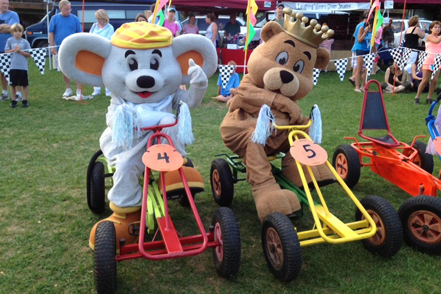 Mouse and bear mascots on go-karts at an outdoor event.