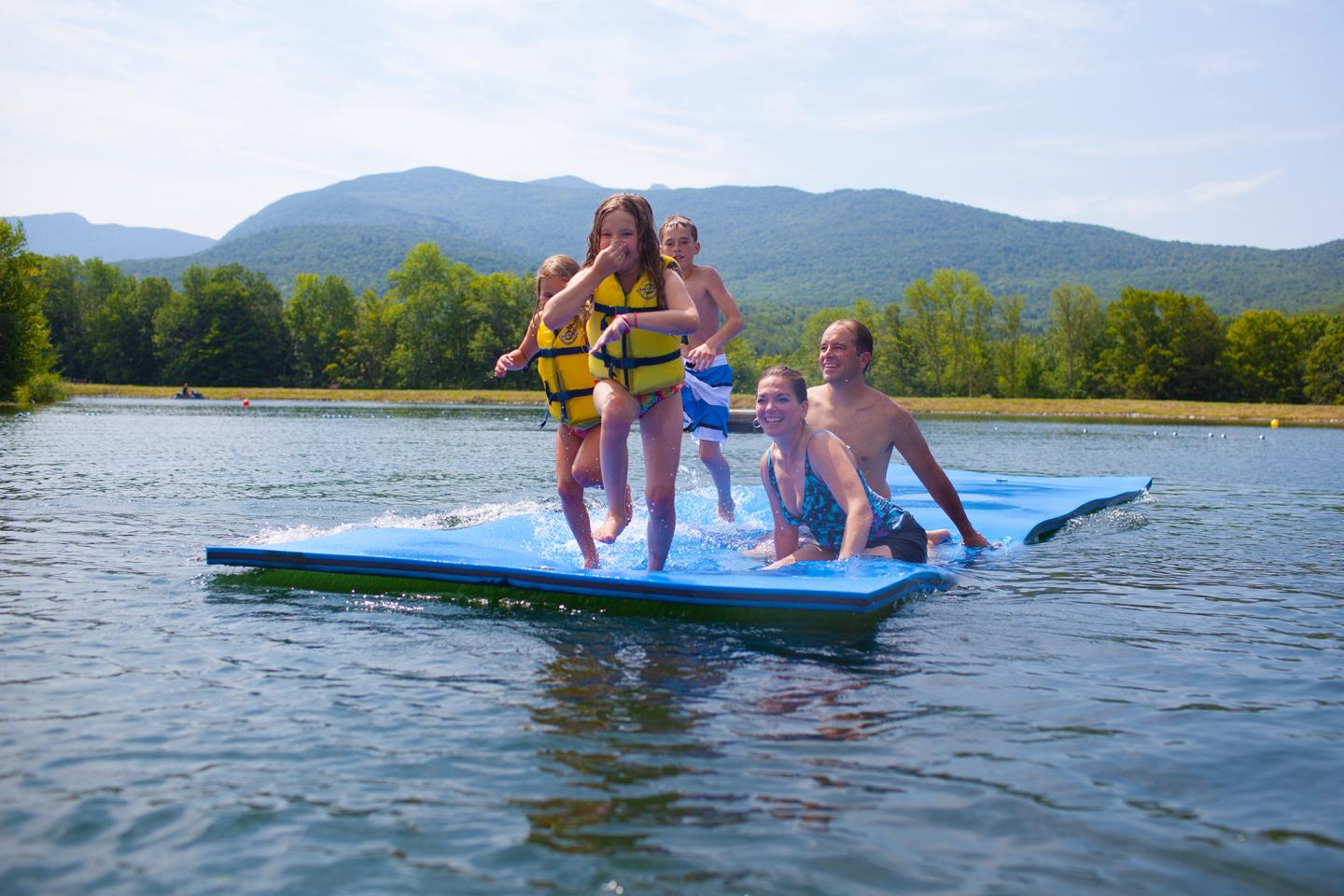 Kids and an adult playing on floating mats in a lake, with mountains in the background.