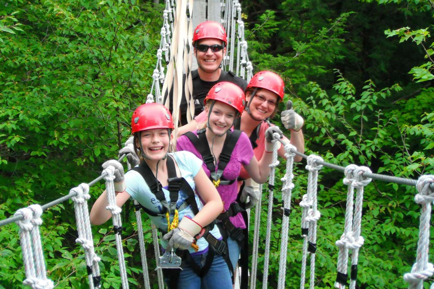 Four people in red helmets smiling on a rope bridge in a forest.
