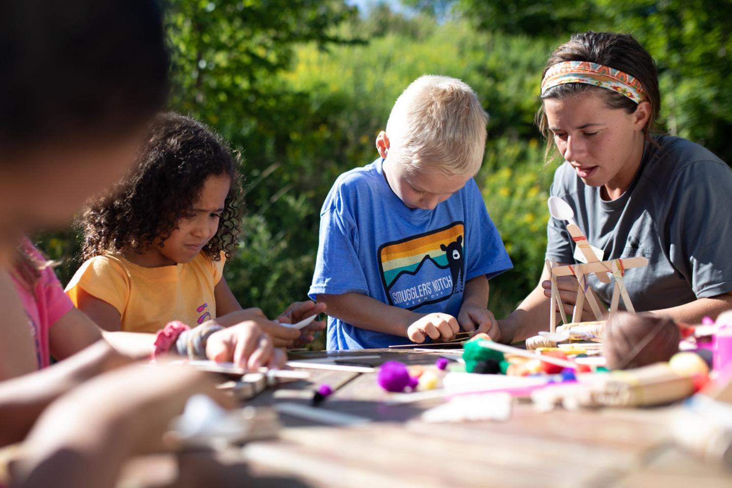 Children crafting outside with an adult, surrounded by colorful materials.