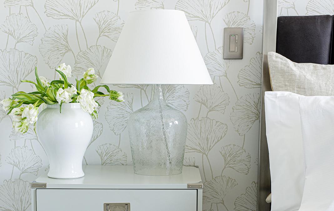White flowers and lamp on bedside table with neutral wallpaper.