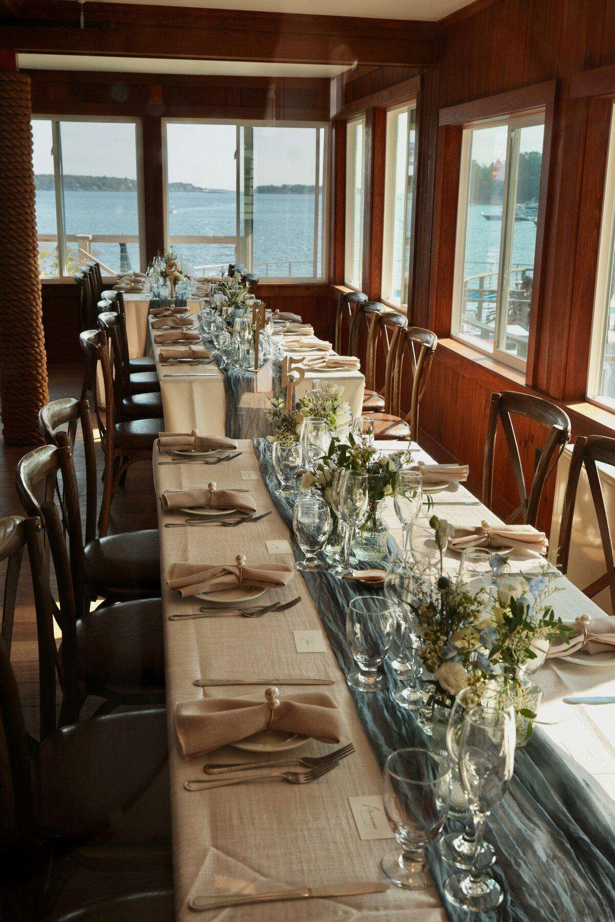 Elegant dining table set for guests with water views through large windows.