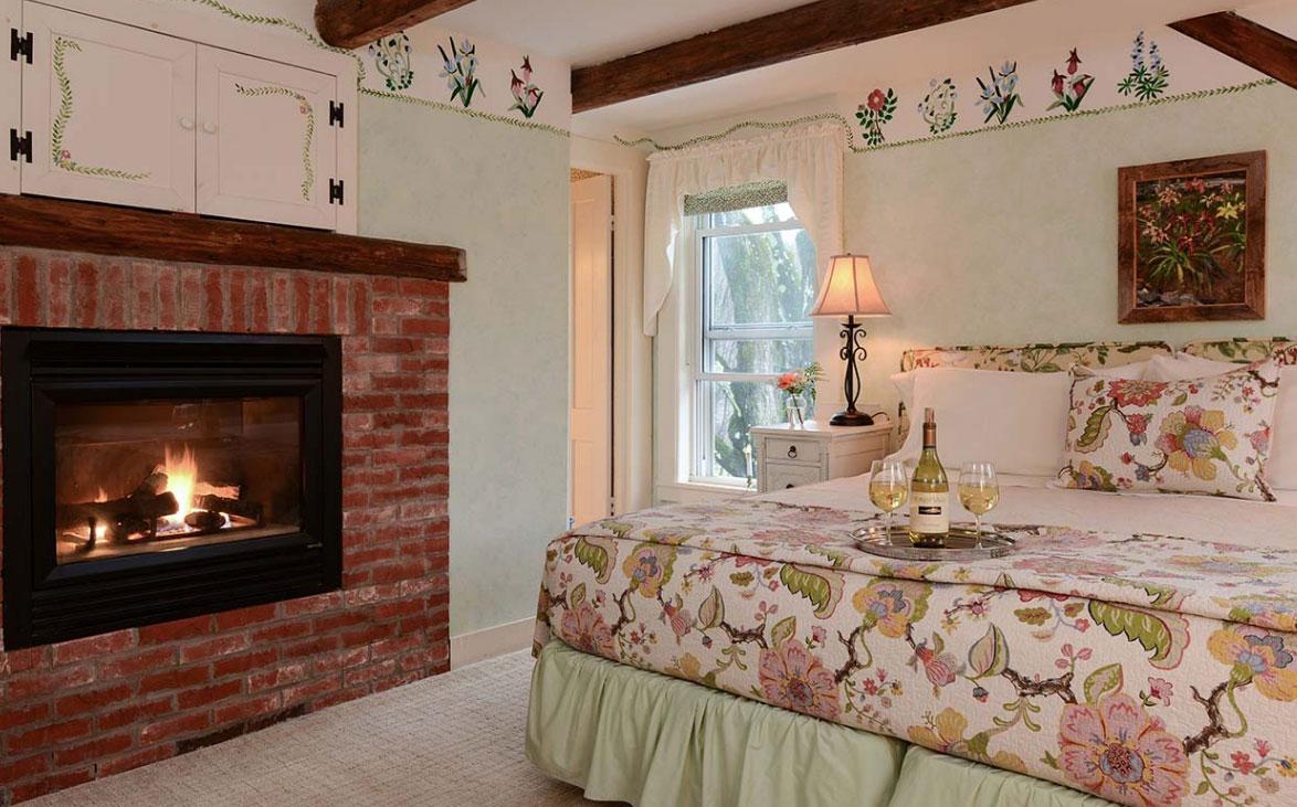 Cozy bedroom with floral decor, lit fireplace, and a wine set on the bed.