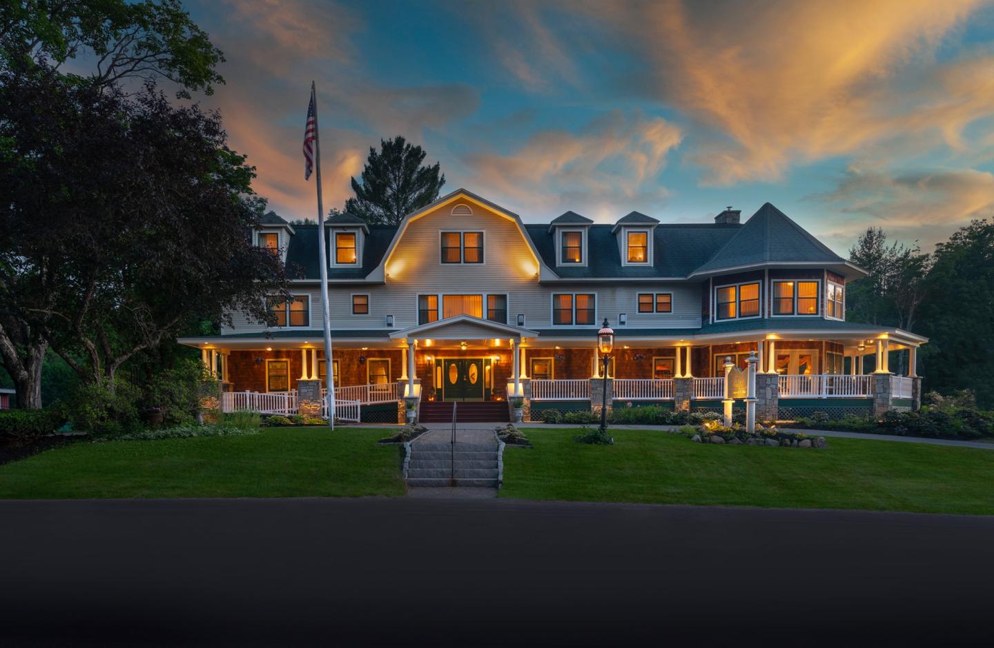 Large house with lit windows at sunset, surrounded by trees.