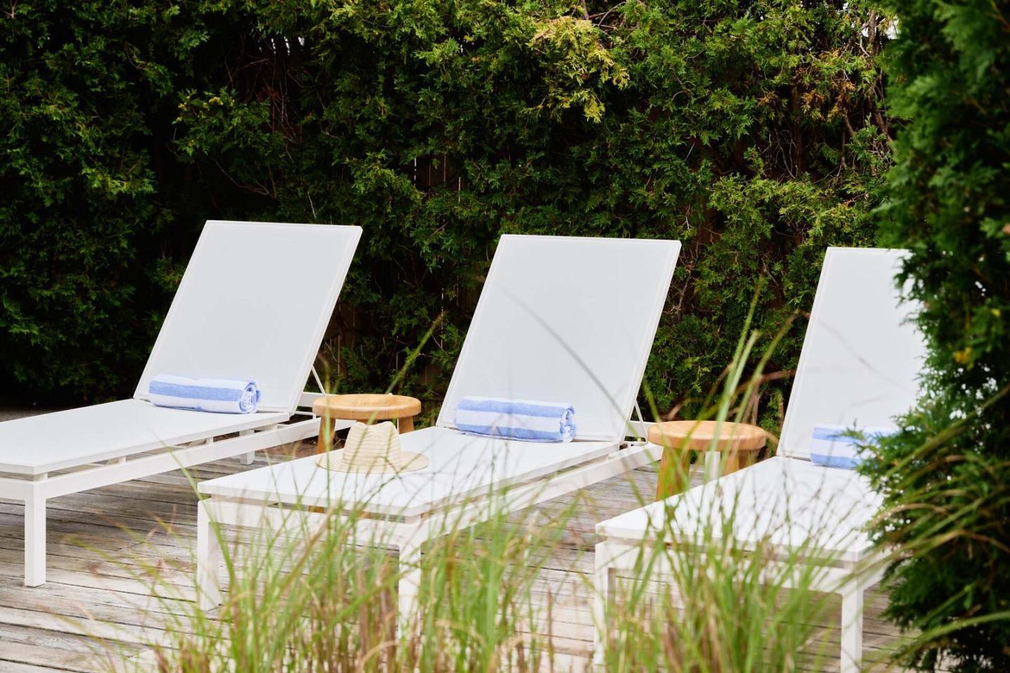 Three white lounge chairs with blue towels on a wooden deck, surrounded by greenery.