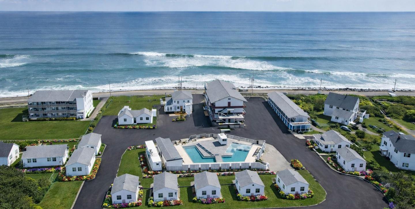Resort with white cottages near ocean, centered pool, lush grass, and crashing waves.