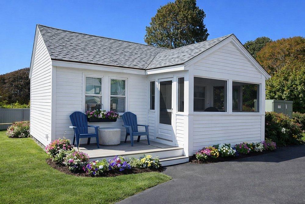 Small white house with a porch, two blue chairs, and colorful flowers.