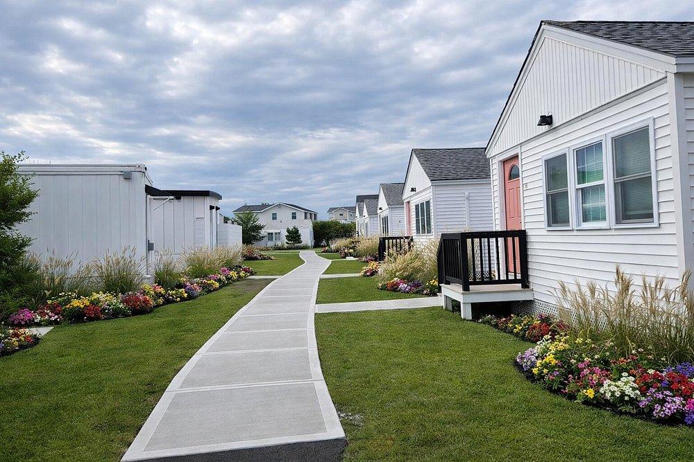 Wide path between white houses with colorful flower beds under a cloudy sky.