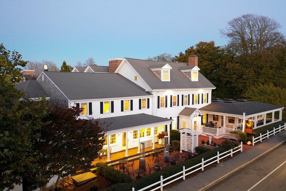 Large white house with lit windows, surrounded by trees, adjacent to a road.
