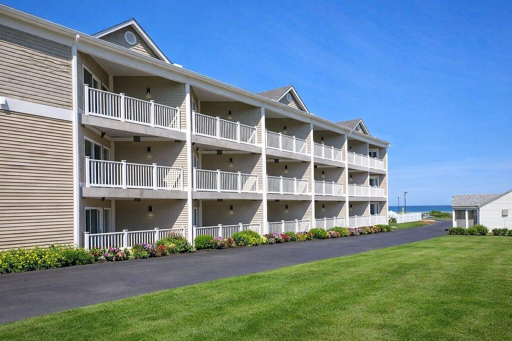 Three-story beige building with balconies, pathway, and grassy lawn under blue sky.