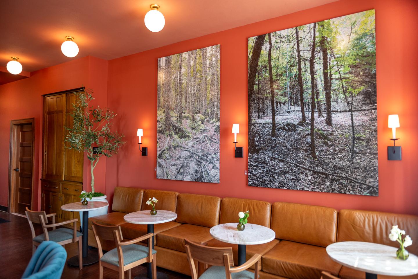 Cozy cafe with orange walls, forest paintings, and round tables with flowers.