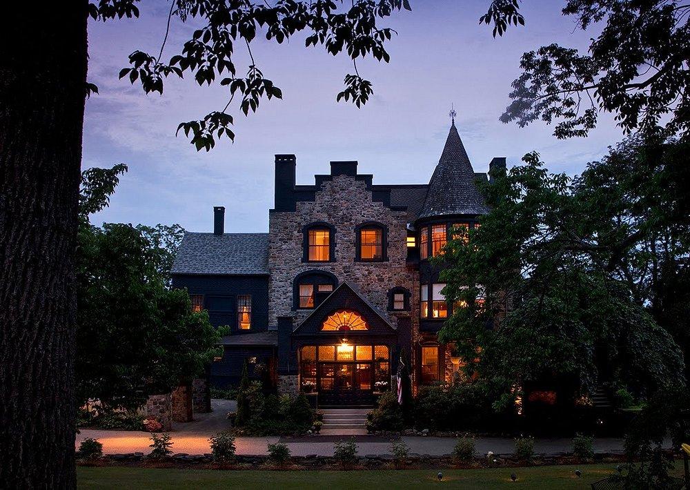 Historic stone building with warm-lit windows at dusk, surrounded by trees.