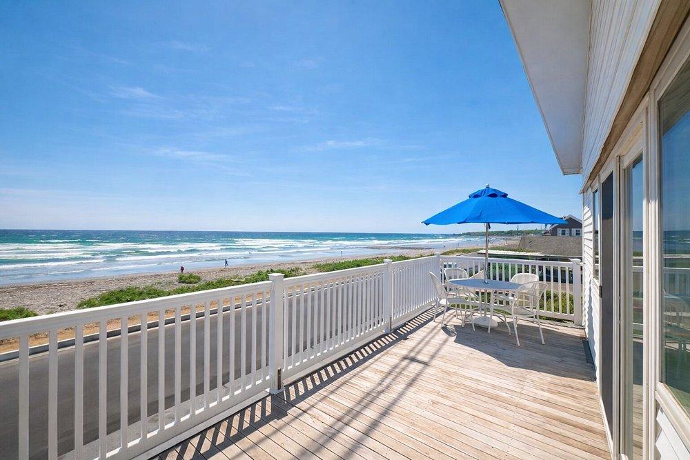 Beachfront deck with white railing, blue umbrella, and ocean view under a clear sky.