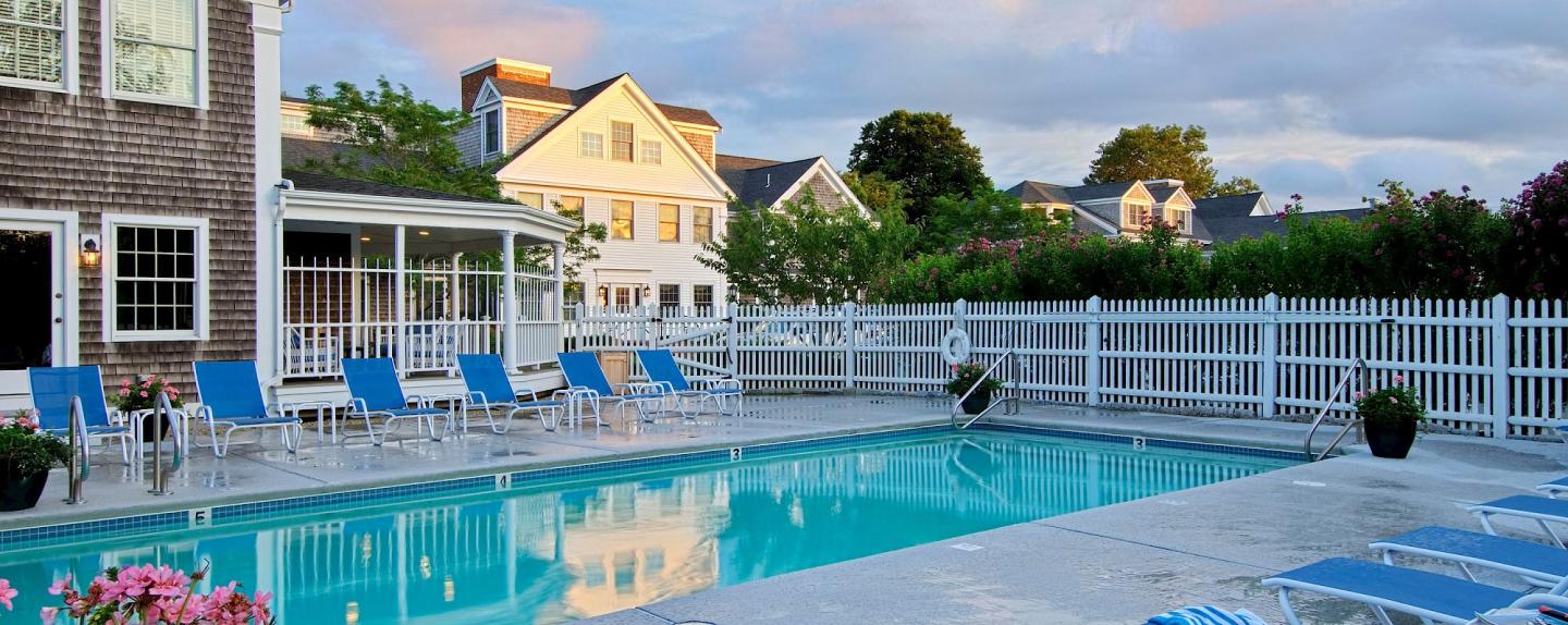 Outdoor pool with blue lounge chairs, surrounded by a white fence and houses.
