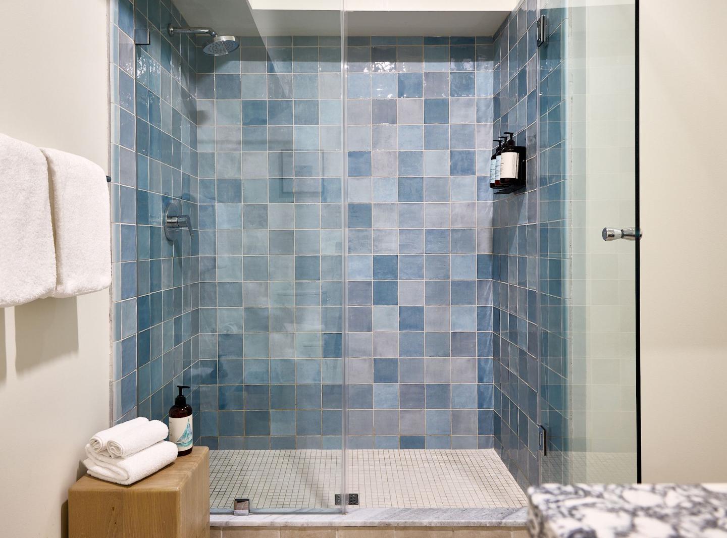 Blue tiled shower with glass door, white towels, and toiletry bottles.