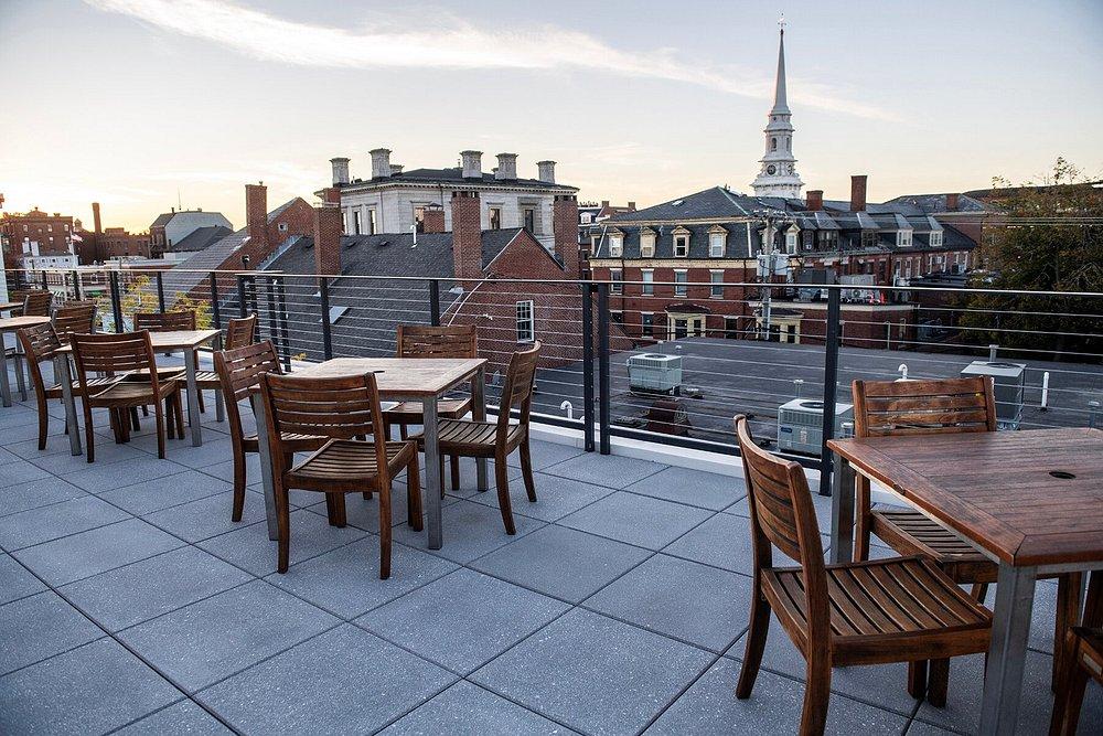 Rooftop patio with wooden tables and chairs, overlooking city buildings at sunset.