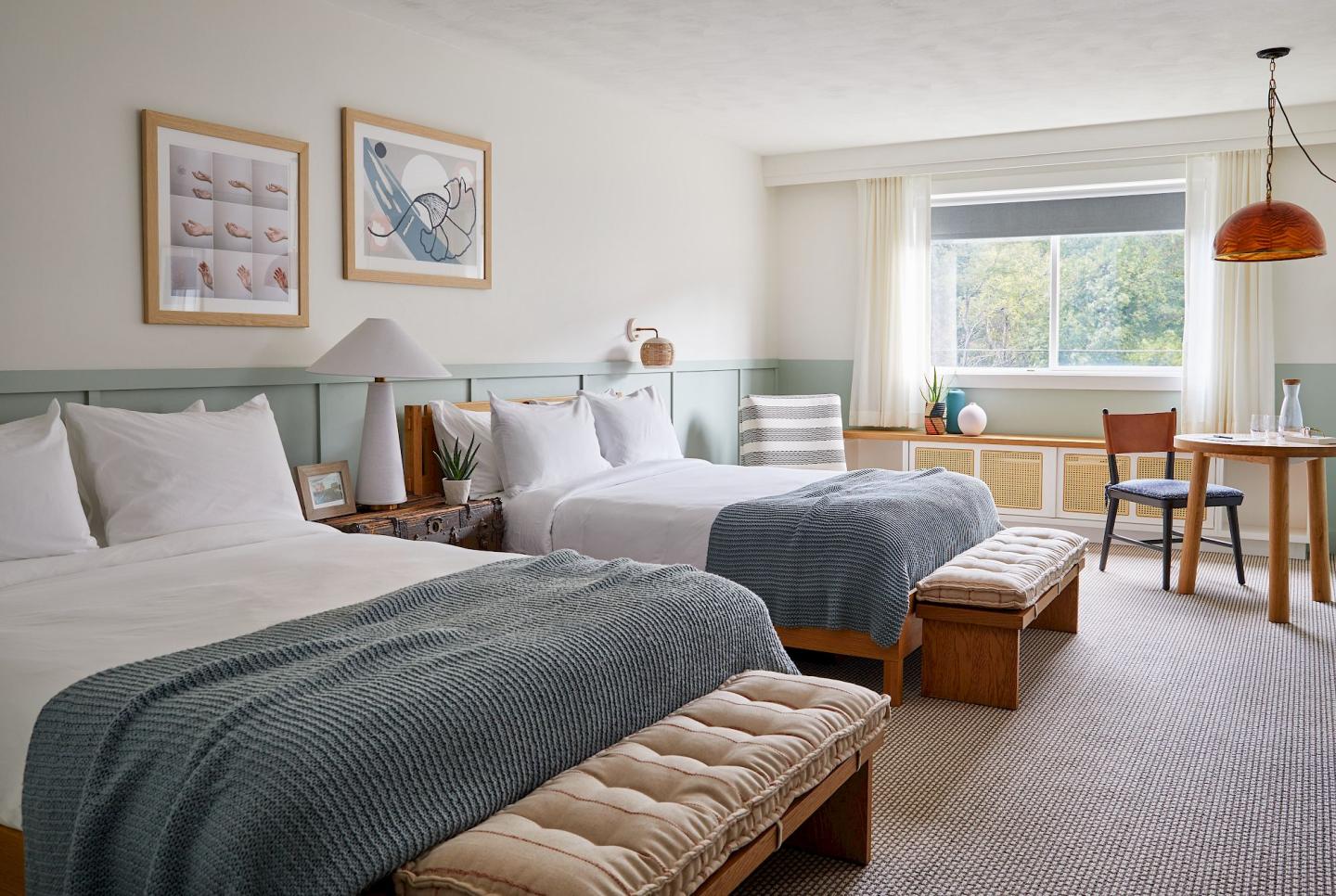 Two neatly made beds in a bright hotel room with a window and desk.