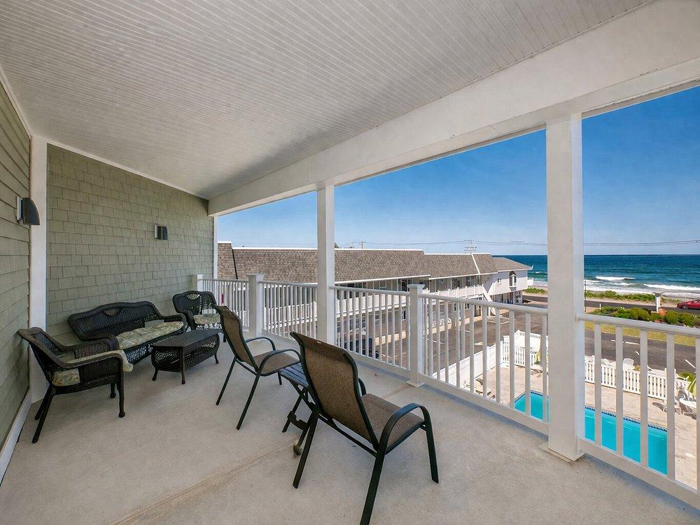 Balcony with chairs overlooking rooftops, pool, and ocean under a clear blue sky.