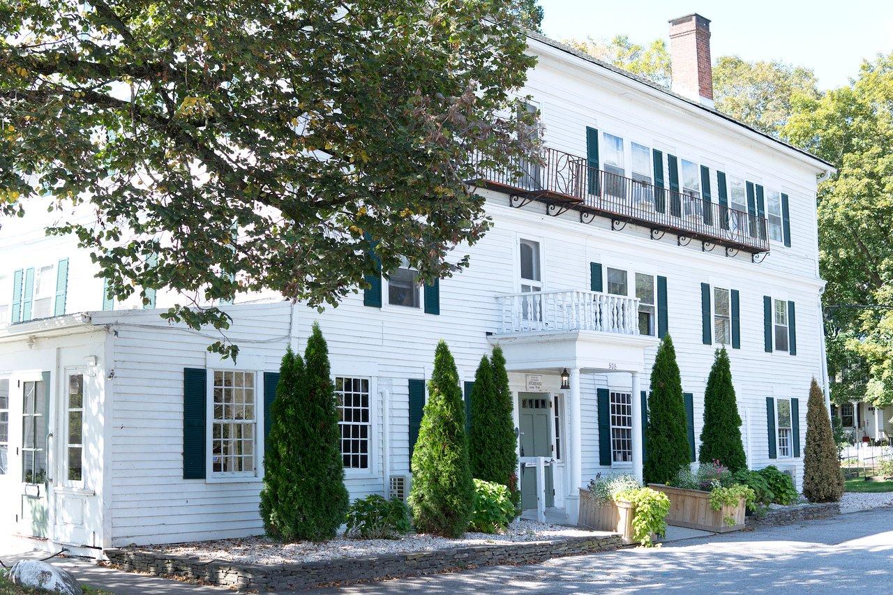 White house with green shutters, trees, and a sunny courtyard.