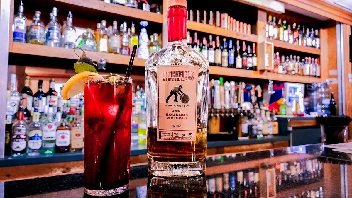 Cocktail and bottle on bar counter with shelves of liquor in background.