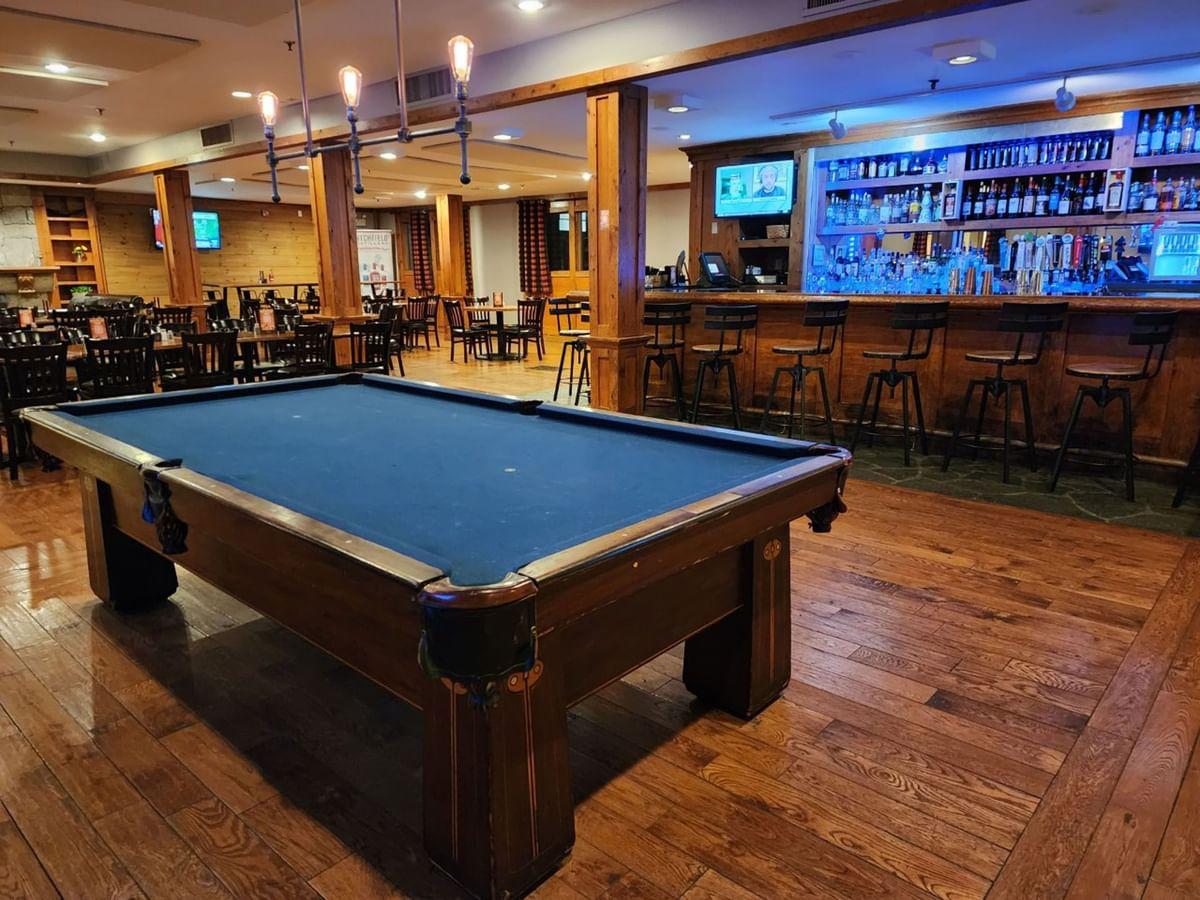 Pool table in a bar with wooden floors and a blue-lit bar area in the background.