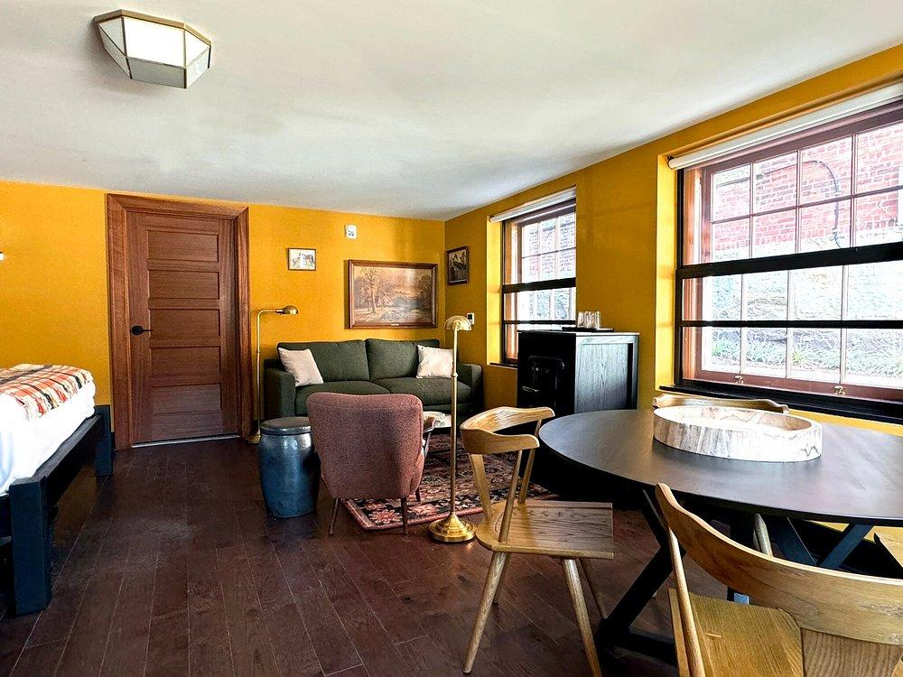 Cozy room with mustard walls, sofa, bed, round table, and large windows.