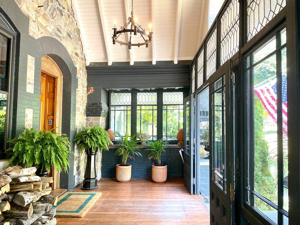 Covered porch with potted ferns, stacked firewood, and an American flag outside.
