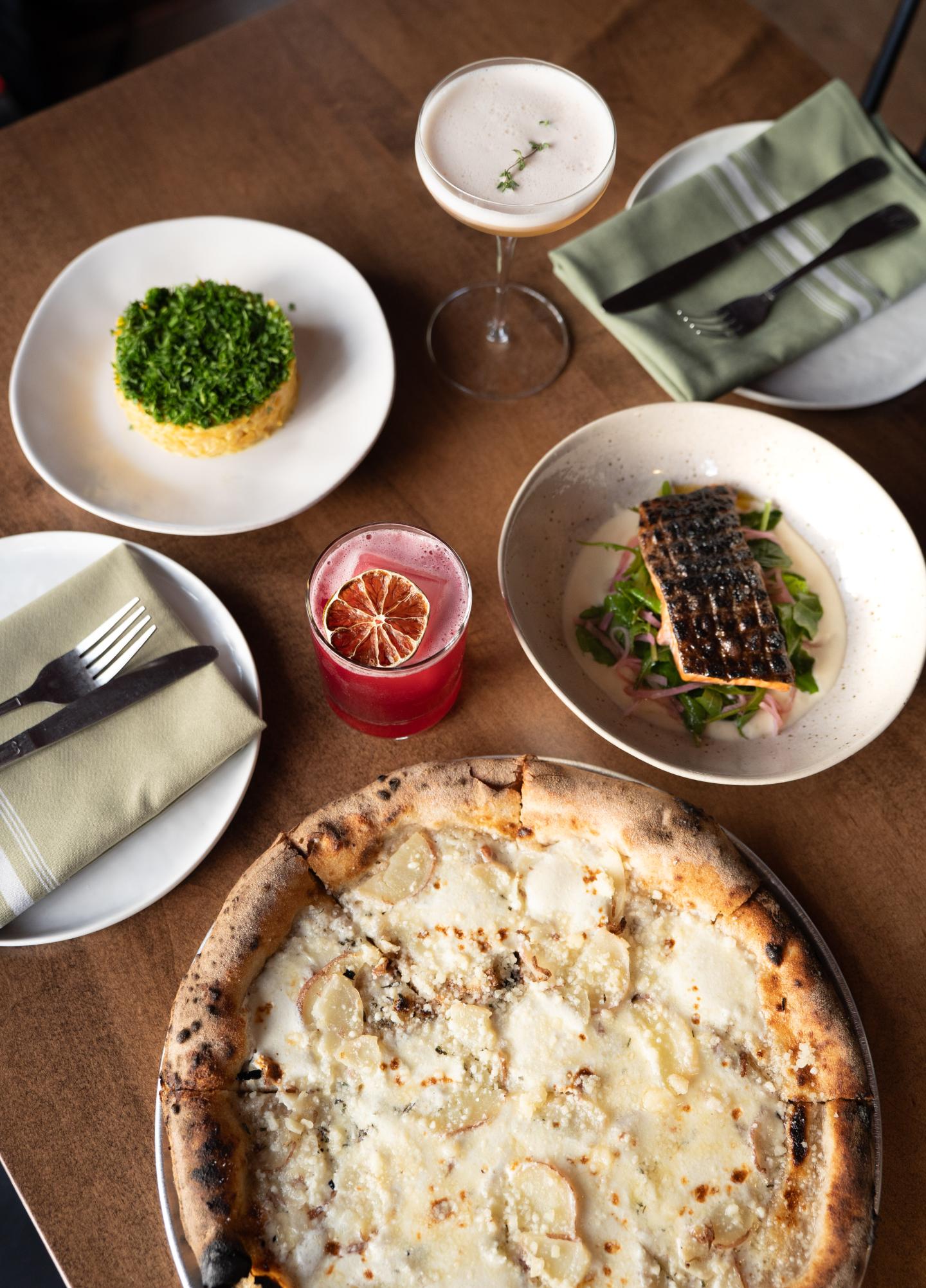 Pizza, drinks, salad, and salmon dish on a wooden table with napkins and cutlery.