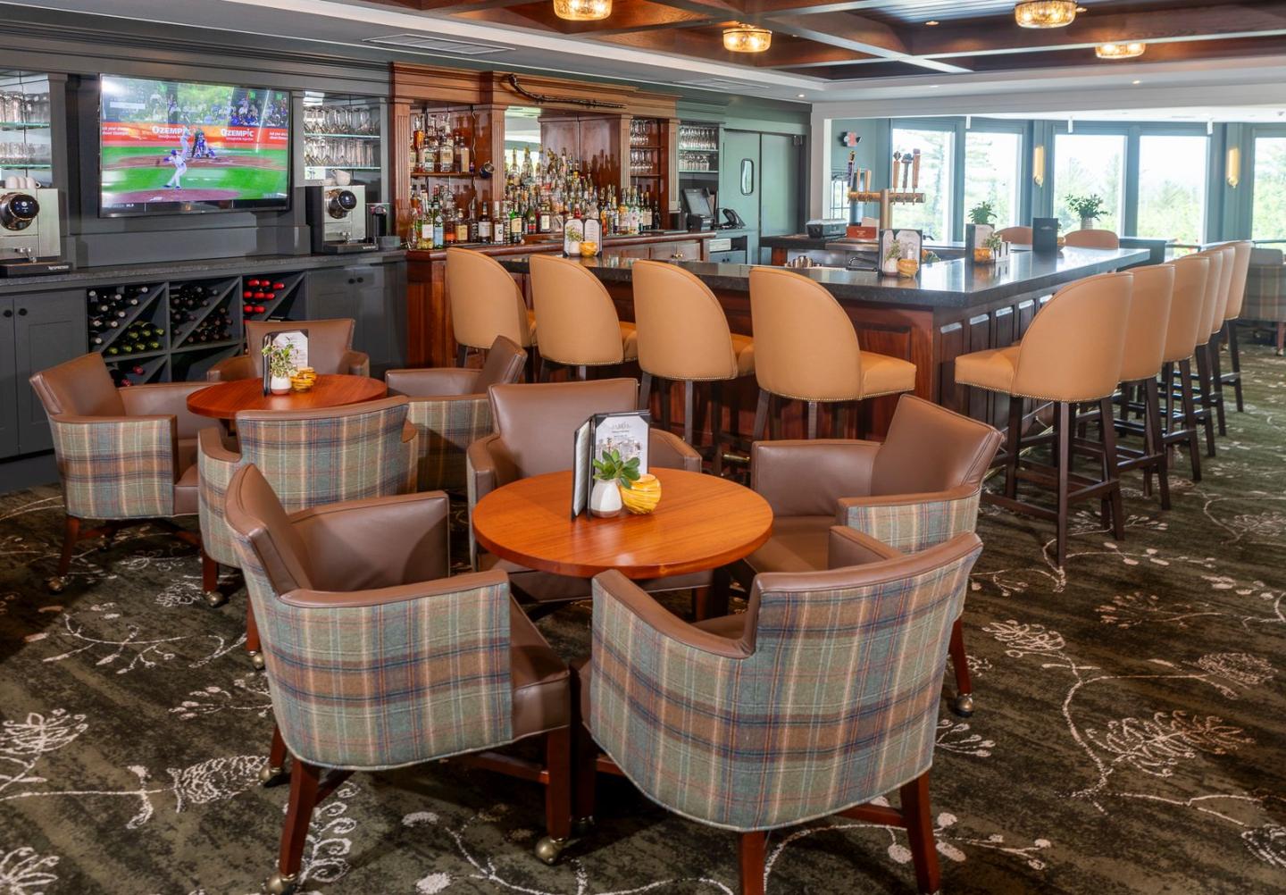 Bar interior with round tables, plaid chairs, and a TV on the wall.