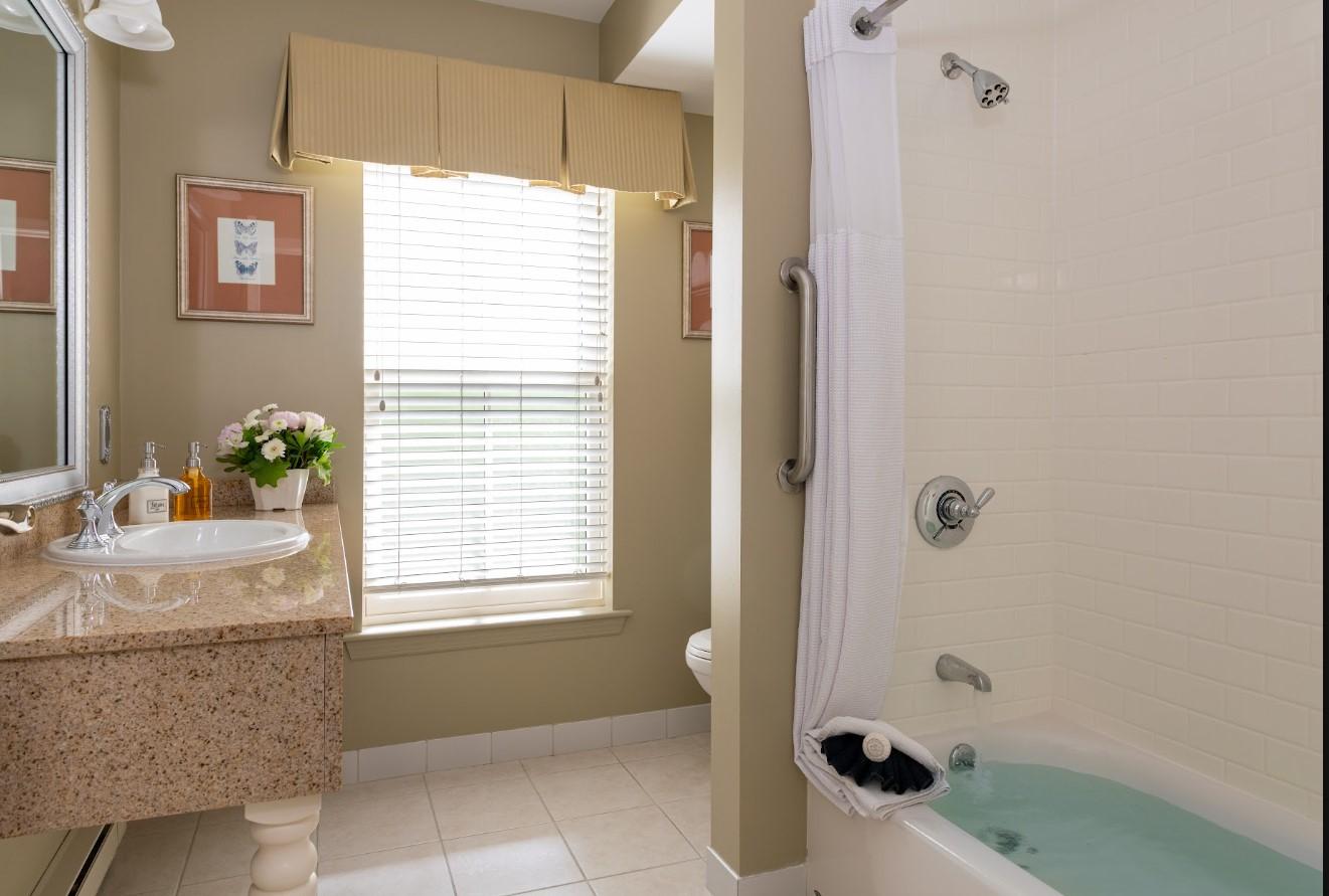 Bathroom with beige walls, granite countertop, flowers, and a bathtub with a safety rail.