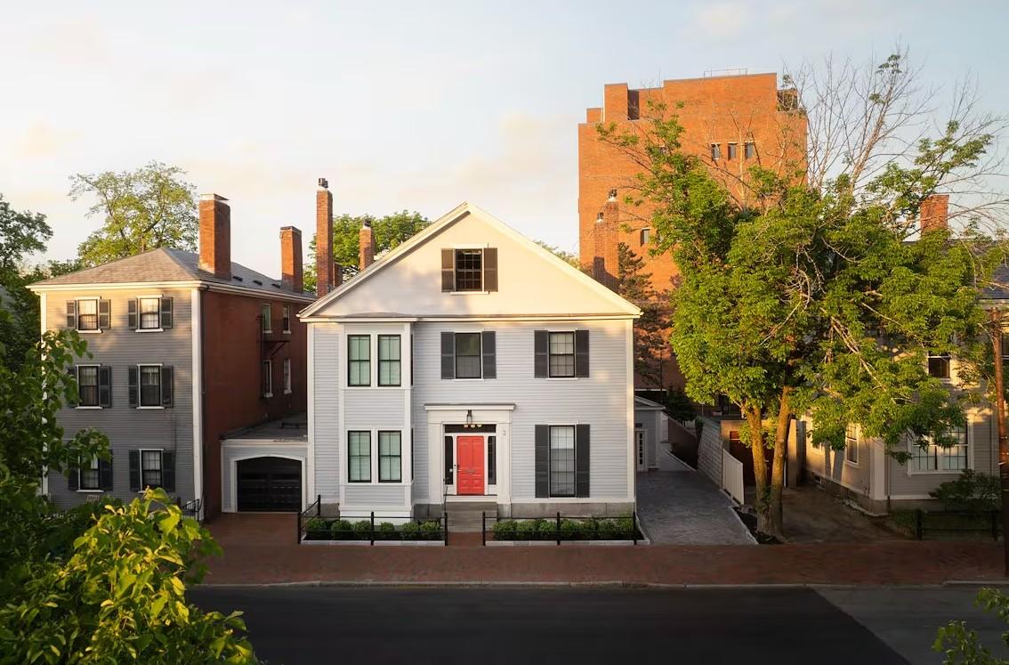 White house with red door, flanked by brick buildings and green trees, under a clear sky.