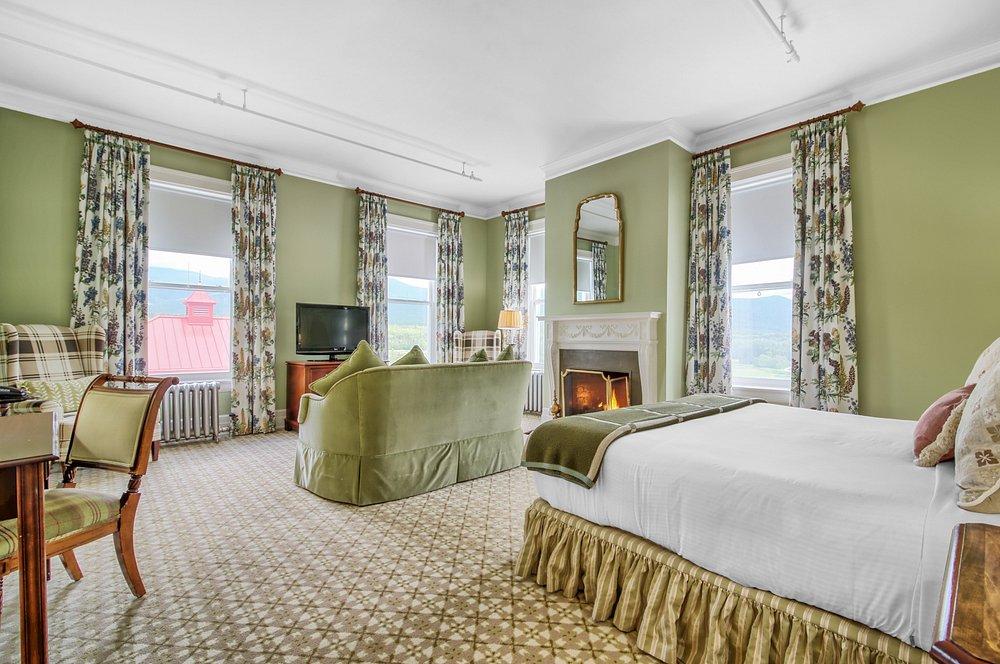 Spacious hotel room with green walls, patterned curtains, fireplace, and a large bed.