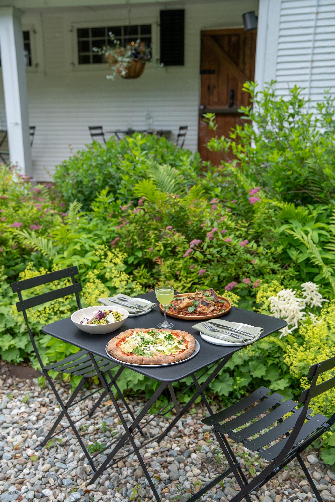 Garden table with pizzas, salad, and drinks by a white house, surrounded by lush greenery.