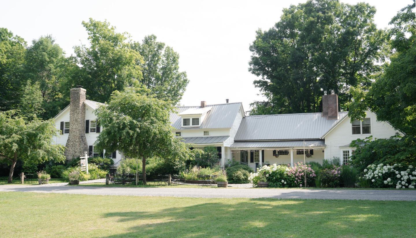 Large white vermont farmhouse inn surrounded by trees and gardens, under a clear sky.