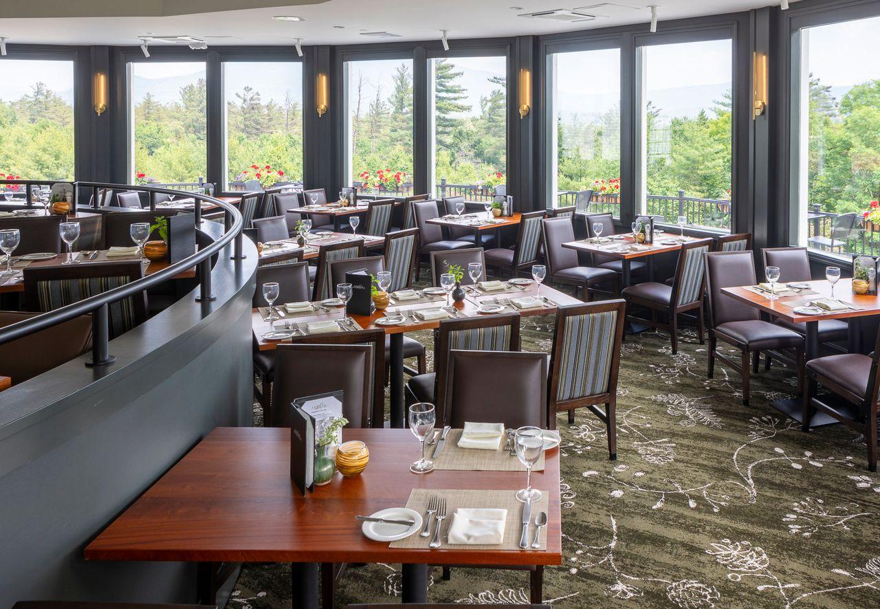 Spacious restaurant with wooden tables, large windows, and lush greenery outside.