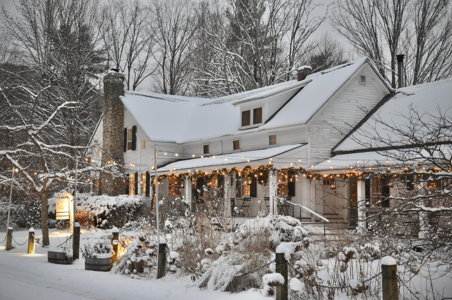Snow-covered Vermont farmhouse inn with festive lights, surrounded by trees.