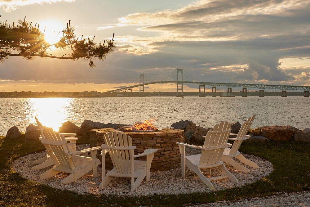 Adirondack chairs around a fire pit by a bay at sunset, bridge in the background.